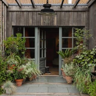 Rustic wooden entryway with open doors, lush potted plants lining the path, and a hanging light overhead.