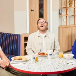 Three friends laugh together at a round table with breakfast plates in a bright cafe setting, cups and juice on the table behind them.