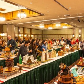 Indoor craft fair with long rows of tables displaying intricate gingerbread houses and people browsing exhibits.",