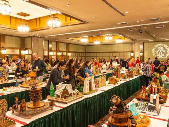 Indoor craft fair with long rows of tables displaying intricate gingerbread houses and people browsing exhibits.",