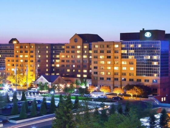 Large brick hotel building at dusk with illuminated windows and blue glass facade, surrounded by trees and a lit parking lot.