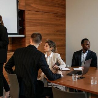 A businesswoman presents at a flip chart to a group seated around a conference table in a modern office.