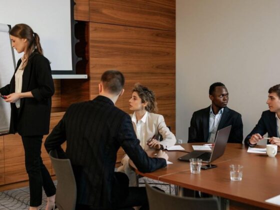 A businesswoman presents at a flip chart to a group seated around a conference table in a modern office.