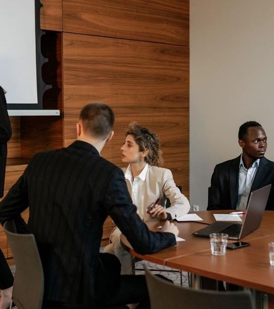 A businesswoman presents at a flip chart to a group seated around a conference table in a modern office.