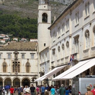 Historic stone town square with arched windows and a tall bell tower, busy with people under white market awnings.