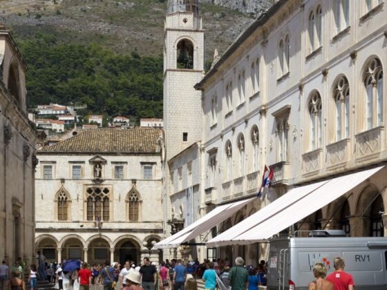 Historic stone town square with arched windows and a tall bell tower, busy with people under white market awnings.