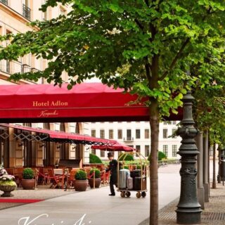 Exterior of Hotel Adlon with a red canopy, outdoor seating, and potted plants along a tree-lined sidewalk, Brandenburg Gate visible in the distance.