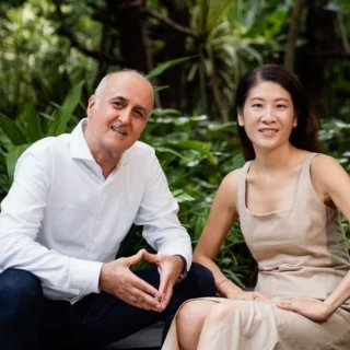 Two adults sit on a low step in a lush garden, smiling at the camera; man wears a white shirt and dark pants, woman a beige dress.