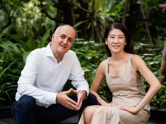 Two adults sit on a low step in a lush garden, smiling at the camera; man wears a white shirt and dark pants, woman a beige dress.