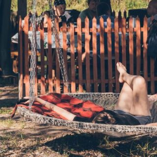Person relaxing on a hanging hammock outdoors with legs raised, a red blanket over them, while onlookers stand behind a wooden fence nearby.