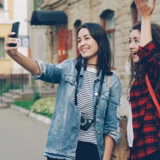 Two young women take a selfie on a sidewalk; one holds a phone, the other raises her hand in the air, both smiling.