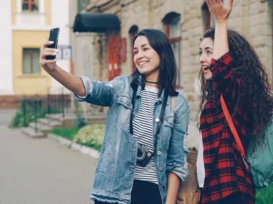 Two young women take a selfie on a sidewalk; one holds a phone, the other raises her hand in the air, both smiling.
