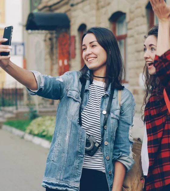 Two young women take a selfie on a sidewalk; one holds a phone, the other raises her hand in the air, both smiling.
