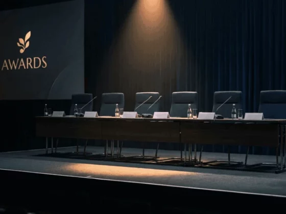Empty awards ceremony stage with a long panel table, seven chairs, microphones and water bottles; screen reads 'AWARDS' on the left.