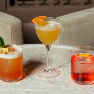 Three colorful cocktails on a round marble table: a peach/orange drink with mint garnish on the left, a yellow cocktail in a stemmed glass in the center, and a red drink with an orange slice on the right, each on a pale scalloped coaster.