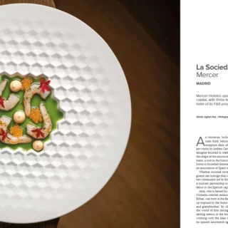 Gourmet appetizer arranged on a textured white plate with a wooden background, next to a magazine page titled La Sociedad Gastronomika Mercer Madrid.