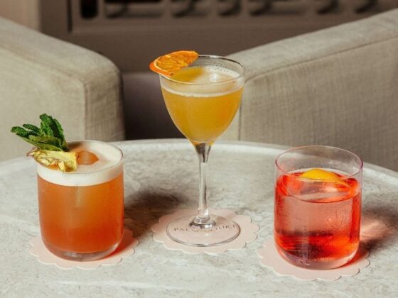 Three colorful cocktails on a round marble table: a peach/orange drink with mint garnish on the left, a yellow cocktail in a stemmed glass in the center, and a red drink with an orange slice on the right, each on a pale scalloped coaster.