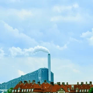 Industrial skyline with a tall chimney emitting white smoke behind red-roofed buildings under a blue sky.