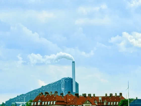 Industrial skyline with a tall chimney emitting white smoke behind red-roofed buildings under a blue sky.
