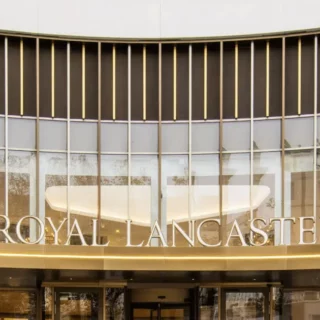 Front entrance of the Royal Lancaster hotel with a curved glass facade and gold accents surrounding the sign 'ROYAL LANCASTER'