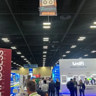Busy convention hall with exhibitor booths, a tall pink banner on the left and a white UniFi booth on the right as attendees mingle.