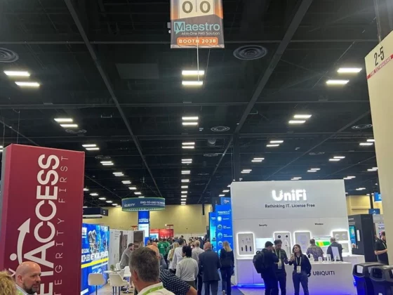 Busy convention hall with exhibitor booths, a tall pink banner on the left and a white UniFi booth on the right as attendees mingle.