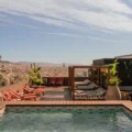 Rooftop pool area with red lounge chairs, wooden deck, cabanas, and potted plants overlooking a city skyline.