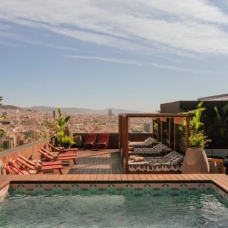 Rooftop pool area with red lounge chairs, wooden deck, cabanas, and potted plants overlooking a city skyline.