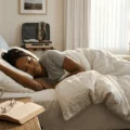 Woman sleeping on a bed in a sunlit hotel room, with an open book, glasses, a coffee mug, and a suitcase in the background.