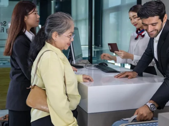 Front desk scene: an elderly guest with a tan purse checks in while a smiling man in a suit assists; two female staff members help nearby on a modern counter.
