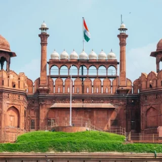 Red sandstone gateway with domed pavilions and multiple arches, topped by an Indian flag against a blue sky (historic monument copy)