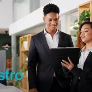 Two business professionals in suits stand at a reception desk, looking at a tablet together in a bright, plant-filled office.