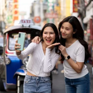 Two young women take a selfie on a busy urban street; one holds a phone while the other smiles, with a blue taxi behind them.