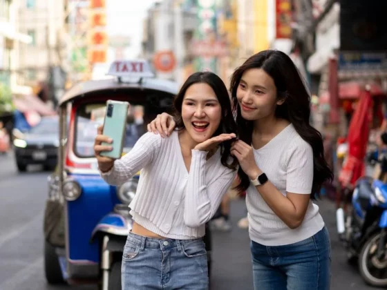 Two young women take a selfie on a busy urban street; one holds a phone while the other smiles, with a blue taxi behind them.