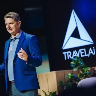 Speaker in a blue suit presenting on stage with a TravelAI logo in the background.