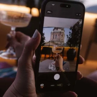 Hand holding a smartphone to photograph a wine glass on a table with a city building in the background viewable on screen.
