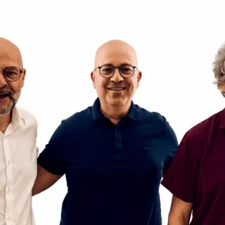 Three men standing side by side, smiling at the camera, wearing white, navy, and burgundy shirts against a white background.