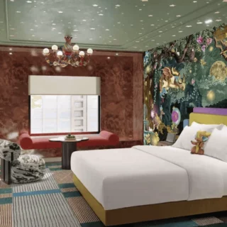 King bed with white linens and a yellow headboard beside a vibrant jungle mural wall and a cozy seating nook with a red sofa and round table.Chic hotel room featuring a colorful mural wall, a king bed, plush seating, and a large window view.Eclectic bedroom with a mural wall, chandelier, and two seating areas: a red sofa and patterned chairs by a window.Bedroom with bold artwork, modern chandelier, and a mixed-material seating area around a small round table.Luxurious hotel room highlighting the vivid wall art, king bed, and a bright, inviting sitting area.