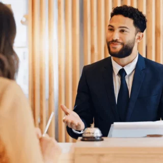 Bearded man in a navy suit at a reception counter, talking and gesturing to a guest.