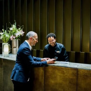 Two men at a wooden hotel reception desk; one in a blue suit shows his phone to the smiling staff member behind the counter.