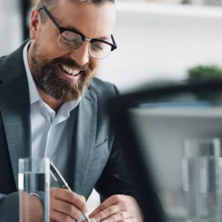 Smiling man in a suit and glasses writes on a document at a modern office desk with a laptop nearby.