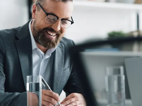 Smiling man in a suit and glasses writes on a document at a modern office desk with a laptop nearby.
