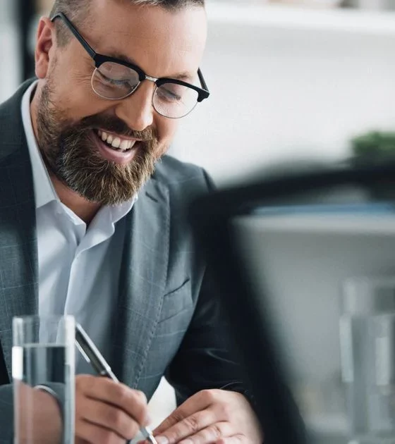 Smiling man in a suit and glasses writes on a document at a modern office desk with a laptop nearby.