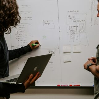Two people brainstorming in front of a whiteboard filled with sketches and sticky notes; one holds a laptop, the other a marker.