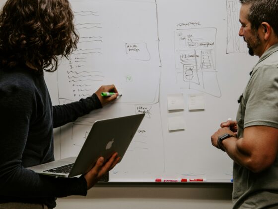 Two people brainstorming in front of a whiteboard filled with sketches and sticky notes; one holds a laptop, the other a marker.