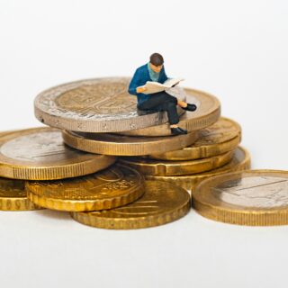 Miniature man sitting on a stack of coins, reading a newspaper, symbolizing finance or savings.