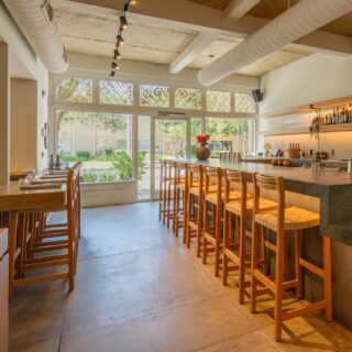 Bright bar interior with a long wooden counter, high stools, and bottles on back-lit shelves beside large windows.