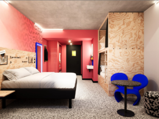 Contemporary hotel room with a low platform bed, pink accent wall, and wood paneling; a small seating area with blue chairs nearby.
