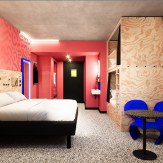 Contemporary hotel room with a low platform bed, pink accent wall, and wood paneling; a small seating area with blue chairs nearby.