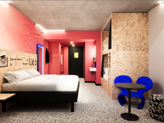 Contemporary hotel room with a low platform bed, pink accent wall, and wood paneling; a small seating area with blue chairs nearby.
